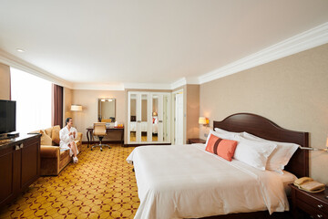 Woman in bathrobe sitting on sofa and drinking coffee in large hotel room with double size bed