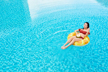 Young Asian woman in red swimwear sitting on inflatable ring and drinking coconut water in swimming pool