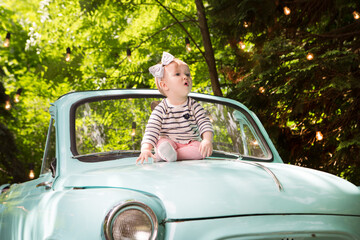 Funny little baby girl sitting on the hood of a car on a sunny day.
