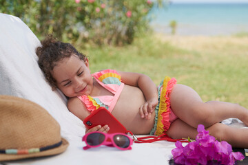 Blonde little girl in pink swimsuit having fun by the sea with mobile phone