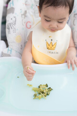 Top view of cute Asian baby girl wearing  baby apron sitting on dining table eat broccoli by her self, Baby Led Weaning concept