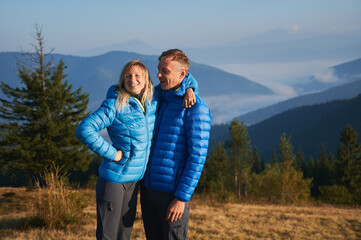 Two happy hikers man and woman standing and embracing on the background of trees, mountain hills, covered by forests and silhouettes mountain peaks far away. Active hiking in the mountains.