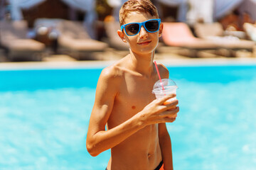 Portrait of a happy boy in sunglasses having a drink near the pool, child with a fresh drink in a...