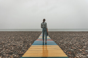 A lonely young woman with bottle of wine walking on empty beach in the rain.