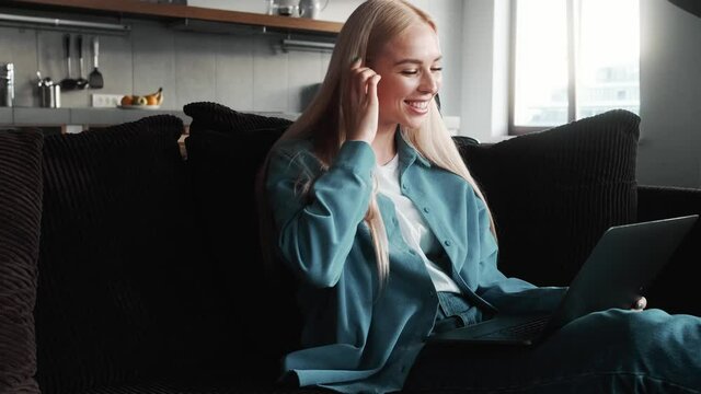 A happy blonde woman is working while talking with video connection on her laptop sitting on the sofa inside the apartments