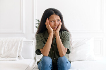 Scared indian woman feels panic attack, sits on the sofa at home, holding head and looks away with fear face expression. Upset mixed-race lady has problem and depression, has poor health, anxiety