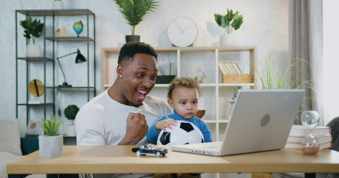 Good-looking happy emotional caring black-skinned dad holding his cute small son on knees when watching football match on laptop and celebrating scored goal,child care and fatherhood concept