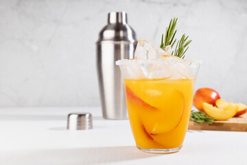 Cooking of homemade organic cold peach smoothie with ice, green rosemary twig, sugar rim, fruit slices in wet glass, ingredients, silver shaker in elegant modern white kitchen interior, closeup.