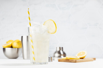 Cold juicy lemon infused cocktail in glass with straw, ice cubes, ingredients in silver bowl, shaker in summer sunlight in soft light white kitchen interior with marble tile.