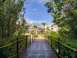 Fototapeta na wymiar View from the wooden bridge surrounded by the jungle to the hotel Tryp Cayo Coco grounds. From the bridge you can see the hotel's white buildings and a seating area with benches for guests.