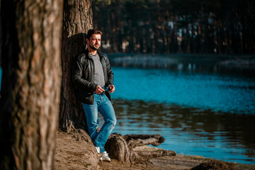 Fototapeta premium Young man in a leather jacket stands pensive and serious under a tree in autumn