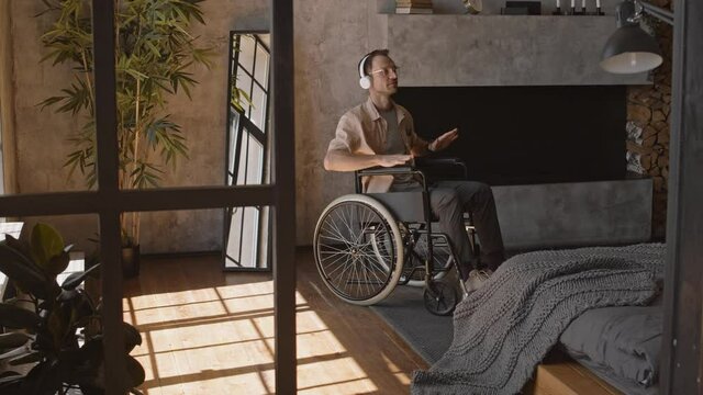 Wide shot of Caucasian man sitting in wheelchair in apartment, wearing eyeglasses and headphones, listening to music using smartphone and enjoying