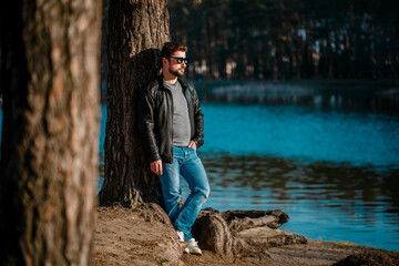 Young man in a leather jacket stands pensive and serious under a tree in autumn