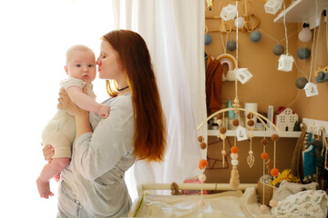Delicate long-haired European mom with a baby 3 months in her arms, mom kisses a baby in a woolen...