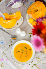 Plate with colorful pumpkin soup, pumpkin cut into pieces on porcelain plates, pepper shaker, salt shaker, spoon, red gerbera flower and green pumpkin seeds  on white tablecloth  