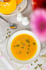 Plate with colorful pumpkin soup, pumpkin cut into pieces on porcelain plates, pepper shaker, salt shaker, spoon, red gerbera flower and green pumpkin seeds  on white tablecloth  