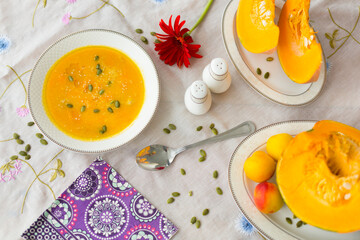 Plate with colorful pumpkin soup, pumpkin cut into pieces on porcelain plates, pepper shaker, salt shaker, spoon, red gerbera flower and green pumpkin seeds  on white tablecloth  
