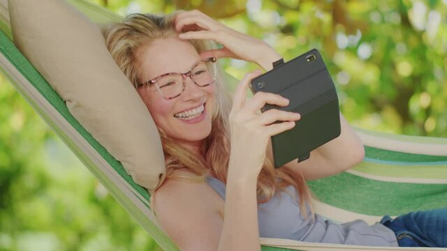 Smiling blonde woman with eyeglasses using smartphone, lying relaxing on the hammock in the garden, free time concept to surf the internet or chat with friends using social media