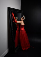 Full length  portrait of beautiful young asian woman wearing red corset, long opera gloves and ornate crown headdress. Graceful posing against a full length mirror with a dark studio background.