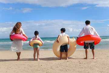 Children with parent wearing colorful inflatable swim ring running to blue sea, kids and adult in a line from behind on tropical sand beach, family spending time  together on summer vacation.
