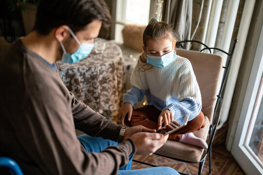 Father with disability in wheelchair using tablet at home with child while wearing masks.