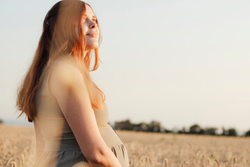 beautiful young pregnant woman walks in ripe wheat field at sunset, expectant mother with red hair relax in nature stroking her belly with her hand