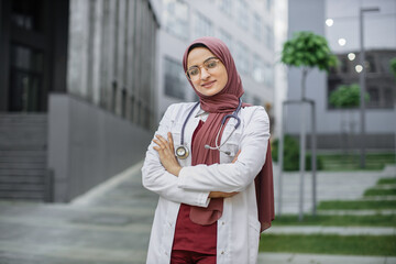 Arab female doctor or medical student, posing and smiling with folded arms, standing outside on a...