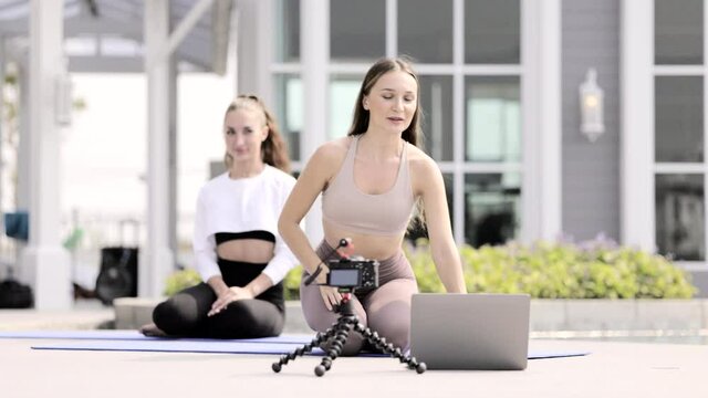 Two beautiful Caucasian women practicing yoga energetic outside of home. Exercise following Pilates video tutorials on laptop. Teaching Workout livestream exercises with camera. sport at home
