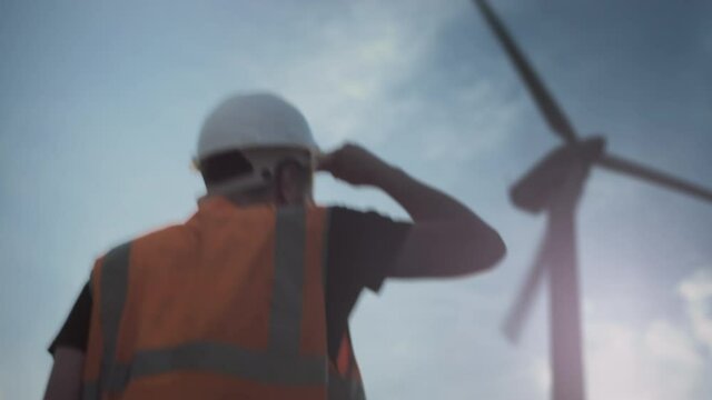 PORTRAIT: A male engineer or worker in a helmet and a vest next to a wind turbine. A young man with a beard, in a white protective helmet and a vest, looks at the camera. Alternative energy sources.