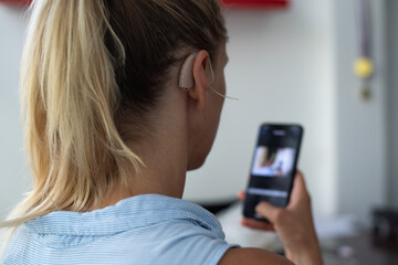 Woman's head in back view, close-up. Person wears beige colour hearing aid on ear and holds phone. Problem with ear. Technology to hear well. Device for communication.