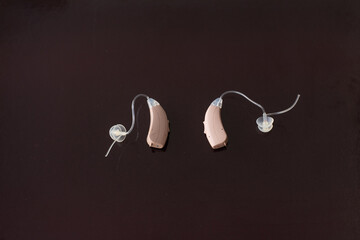 Two beige hearing aids for hearing impairment, different positions, isolated on brown dark background. Device for well communication.