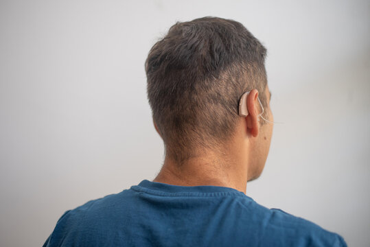 Man's head in back view, close-up. Person wears beige colour hearing aid on ear. Problem with ear. Light background. Technology to hear well. Device for communication.