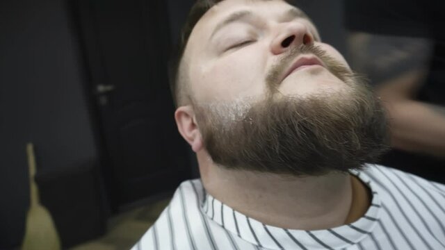 Inside barbershop salon. Barber with stainless steel straight edge shaving razor knife trims beard of plump chubby client. Grooming, haircut and shaving service. Closeup. Caucasian man