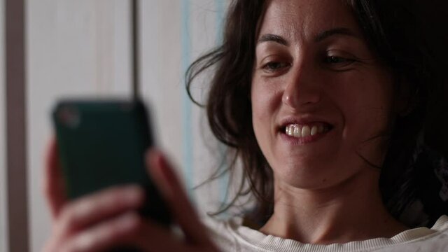 Smiling girl lies on the couch and writes a message on the smartphone, the woman is resting, the brunette reads and smiles