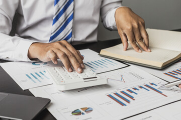 Businessman is pressing a calculator and looking at the information in his notebook, he is checking the balance from the monthly financial and sales documents. Concept of finance and sales management.