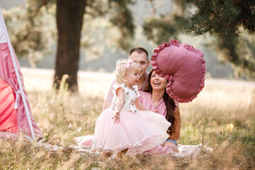 Girl are dressed in pink dress and holding big pink heart in hands. Mom, dad are sitting next to wigwam decoration in the park. Family spending time outdoor in summer, having fun together. © Andriy Medvediuk