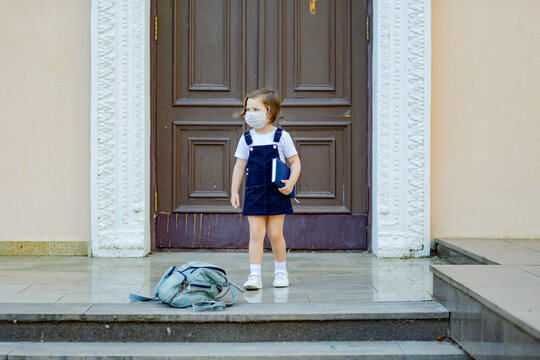 a beautiful little girl, a schoolgirl, stands outside the school during the day, a medical mask on her face