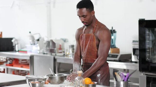 Handsome African American Man Chef Wearing Brown Leather Apron Kneading Dough On The Table. Happy Cooking Black Guy. Making Bread, Manufacturing Process. Workplace, Professional Kitchen Interior.