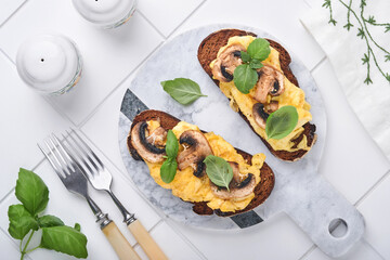 Scrambled eggs with fried mushrooms and basil on bread on white table background. Homemade breakfast or brunch meal. Scrambled eggs and mushrooms sandwiches. Top view with copy space