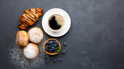 Donuts (buns) with powdered sugar, blueberries, croissant and coffee for breakfast. Black background. Top view with copy space.