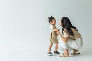 full length of happy asian toddler kid holding hands with stylish mother on grey - Powered by Adobe