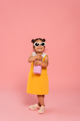full length of cheerful asian toddler kid in yellow dress and sunglasses holding present on pink