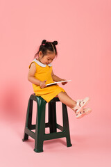 full length of pleased asian toddler kid in yellow dress sitting on chair and using digital tablet on pink