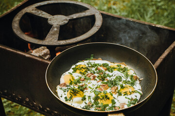 Frying pan of fried eggs, bacon and organic greens, green grass on the background, cooked on the grill on a summer sunny day, summer breakfast, eating in nature - the concept of a healthy lifestyle