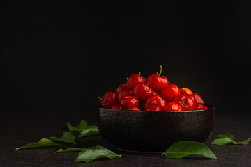 Side view of ripe red acerola cherries fruit in a ceramic bowl with a black background. High vitamin C and antioxidant fruits. Close-up photo. Space for text. Concept of healthy fruits