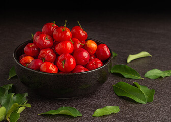Top view of ripe red acerola cherries fruit in a ceramic bowl with green leaves on a stone table. High vitamin C and antioxidant fruits. Close-up. Space for text. Concept of healthy fruits