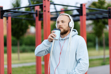 Obraz premium A man with a braid hairstyle in the early morning drinks coffee on the sports ground and listens to music on headphones
