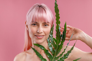 Portrait of beautiful naked woman with pink hair smiling at camera, posing with plant leaf isolated over pink studio background. Beauty, skin care, natural cosmetics concept