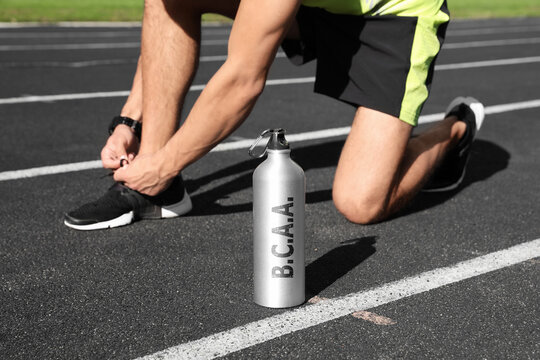 Bottle with amino acids (BCAA) drink and young man tying shoelaces at stadium