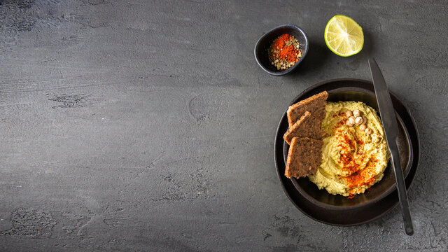 Chickpea hummus  decorated with sweet paprika and bread tosts on dark background with spice bowl,lemon, and knife.Large image for banner